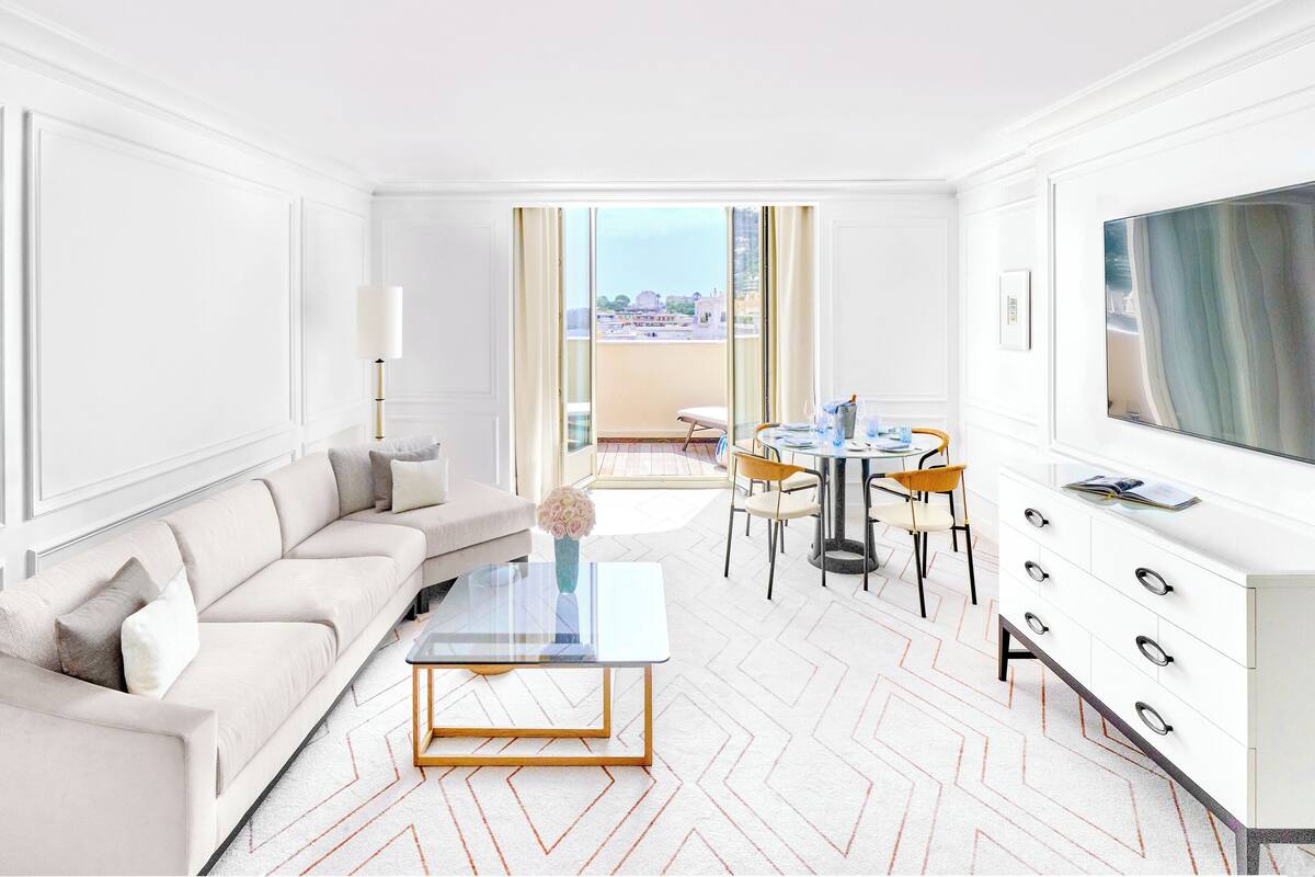 A modern hotel room with a sofa, a glass coffee table, a dining area with chairs, a TV, and a view of the city.