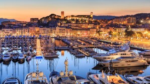 A marina with numerous boats docked, a prominent castle on a hill, and a cityscape with buildings in the background. - Carlton Cannes, a Regent Hotel (Cannes)