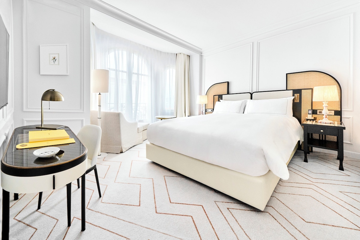 A hotel room with a large bed, a desk with a lamp, a chair, and a nightstand with a lamp.