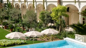 Une piscine agrémentée de trois parasols blancs, entourée de verdure et bordée d’un bâtiment aux fenêtres en arc de cercle.