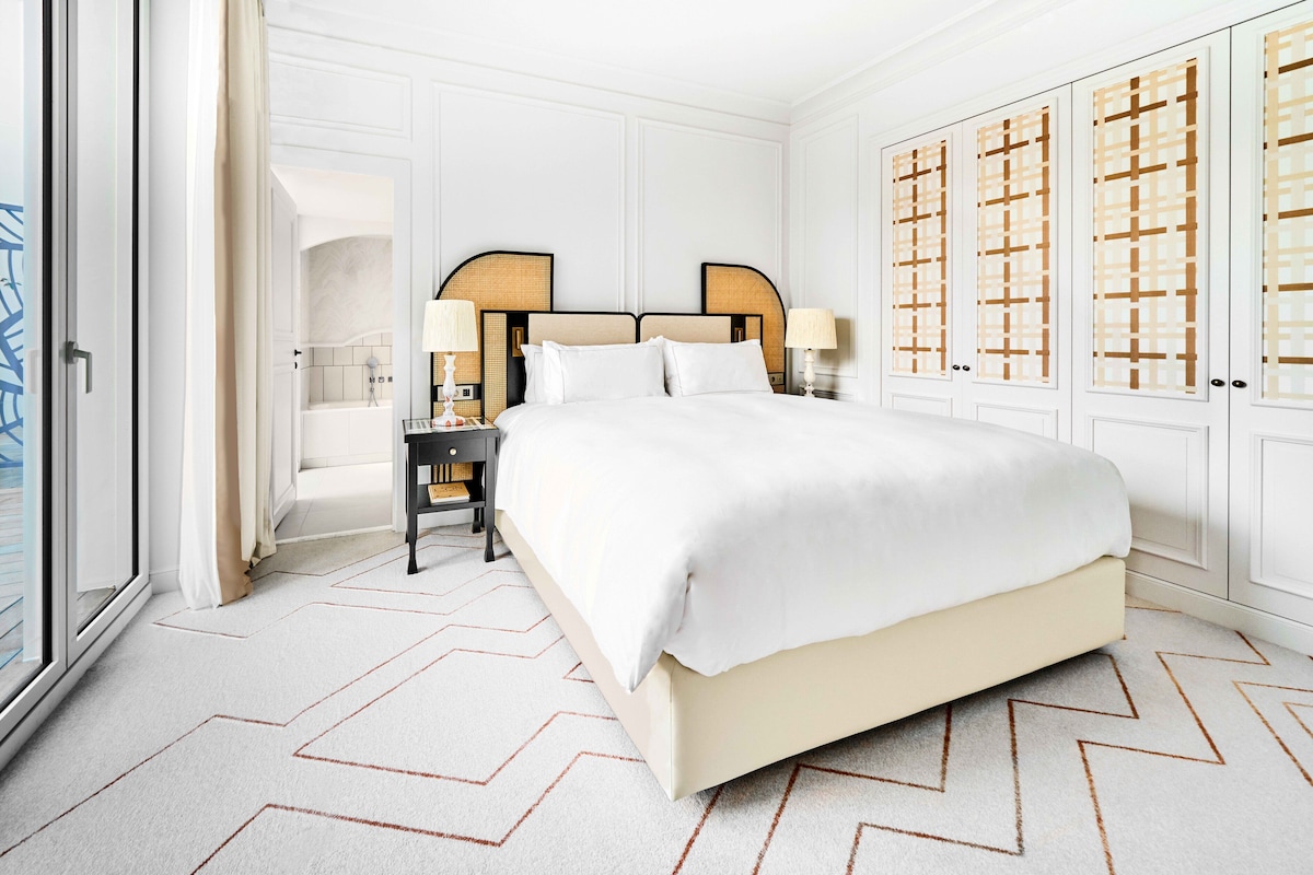 A spacious bedroom with a large bed, a nightstand, a lamp, and a wardrobe with a geometric pattern.