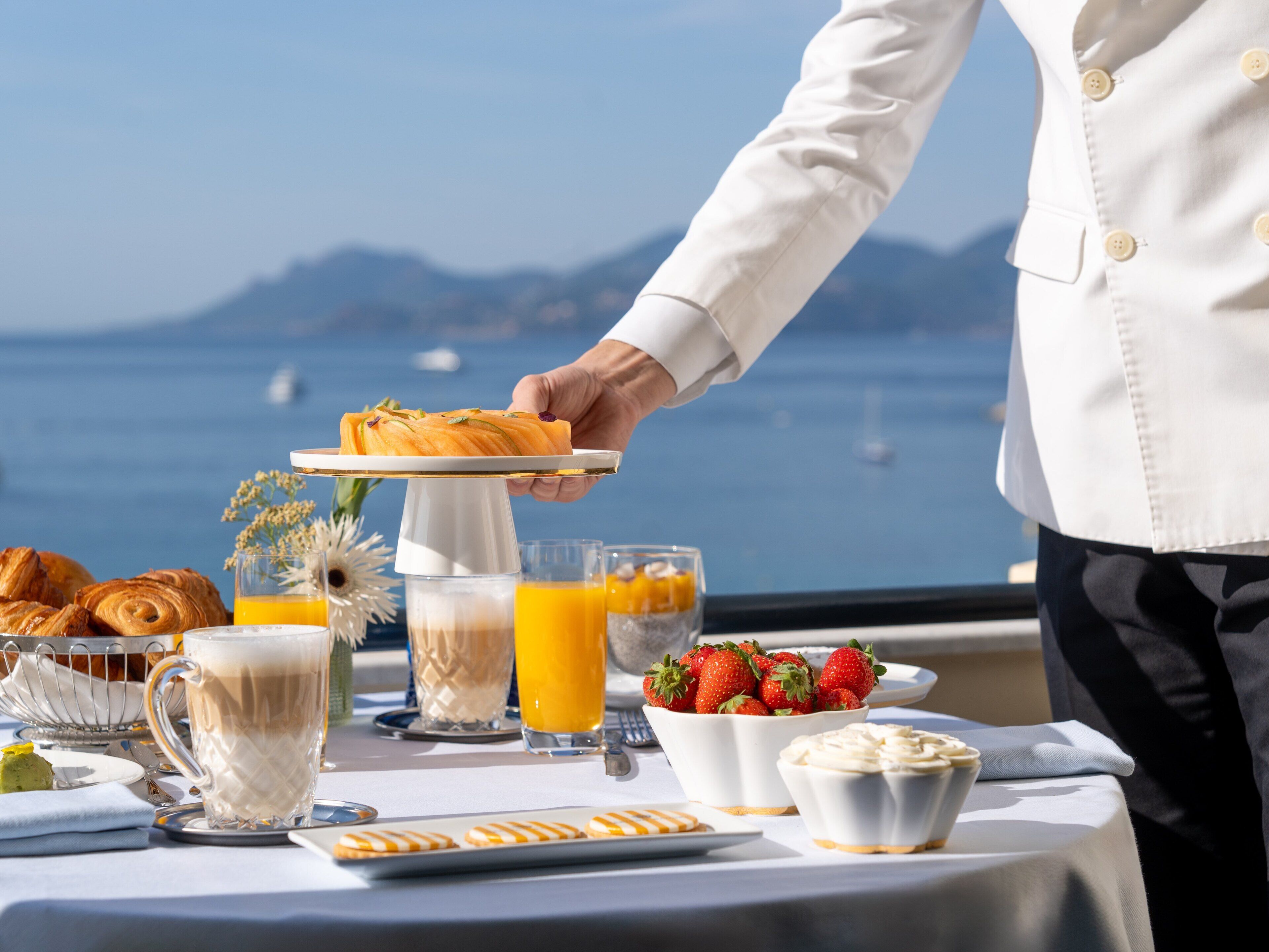 A breakfast setting with croissants, fruit, coffee, and juice on a table with a view of the sea.