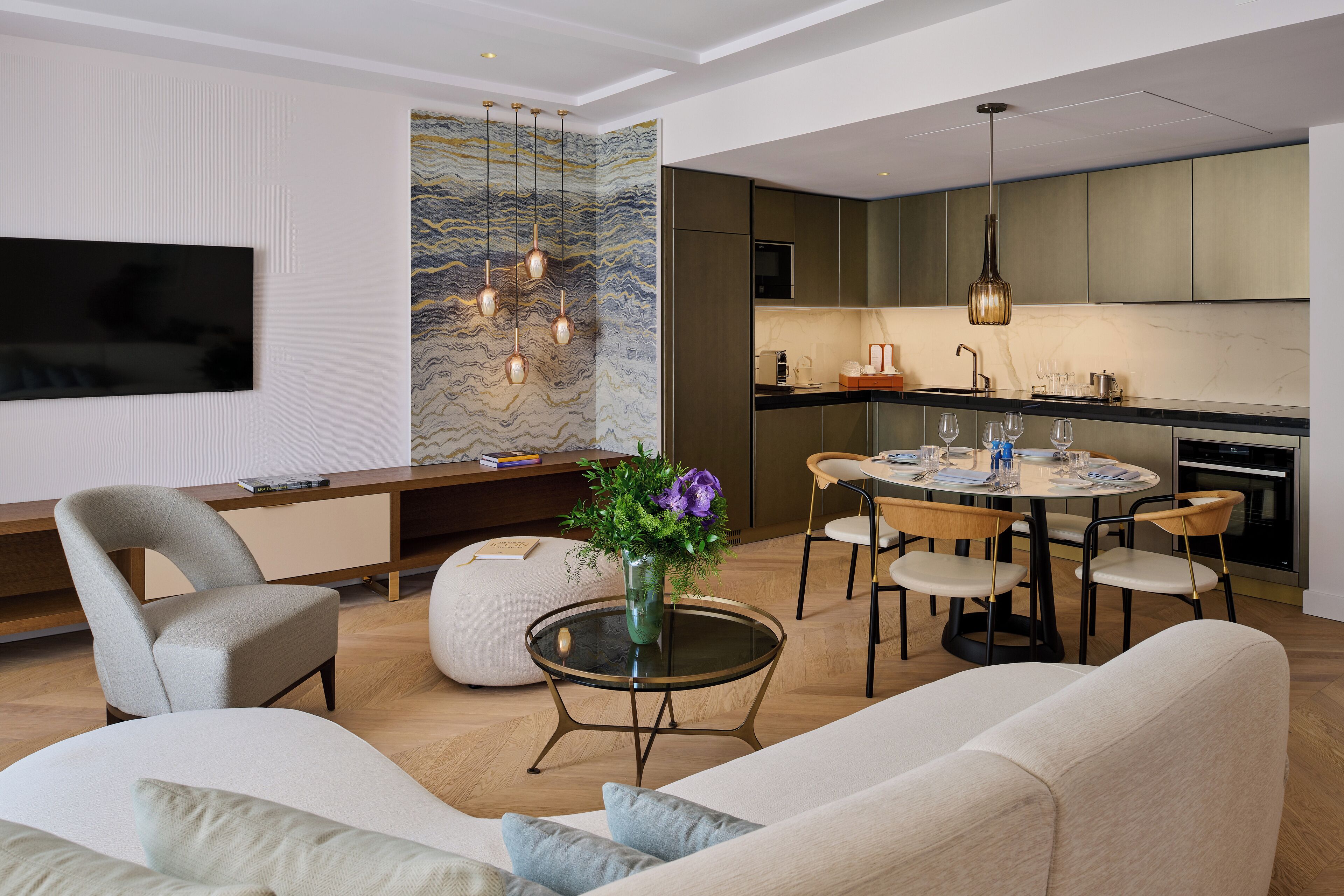 A modern kitchen with a dining area, a flat-screen TV mounted on the wall, and a decorative vase with flowers on the dining table.