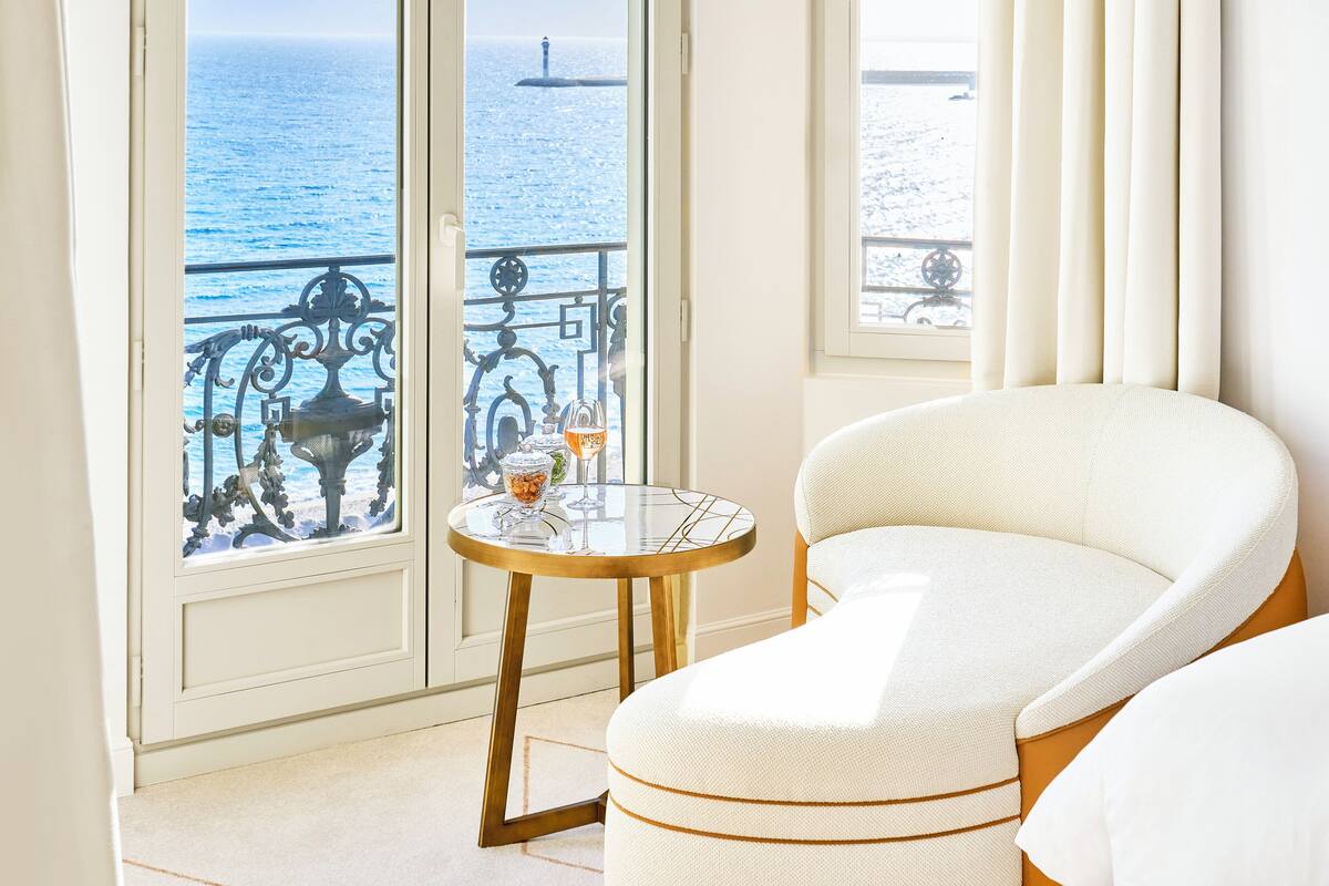 A room with a view of the sea, a round table with a glass top, and a white upholstered chair.