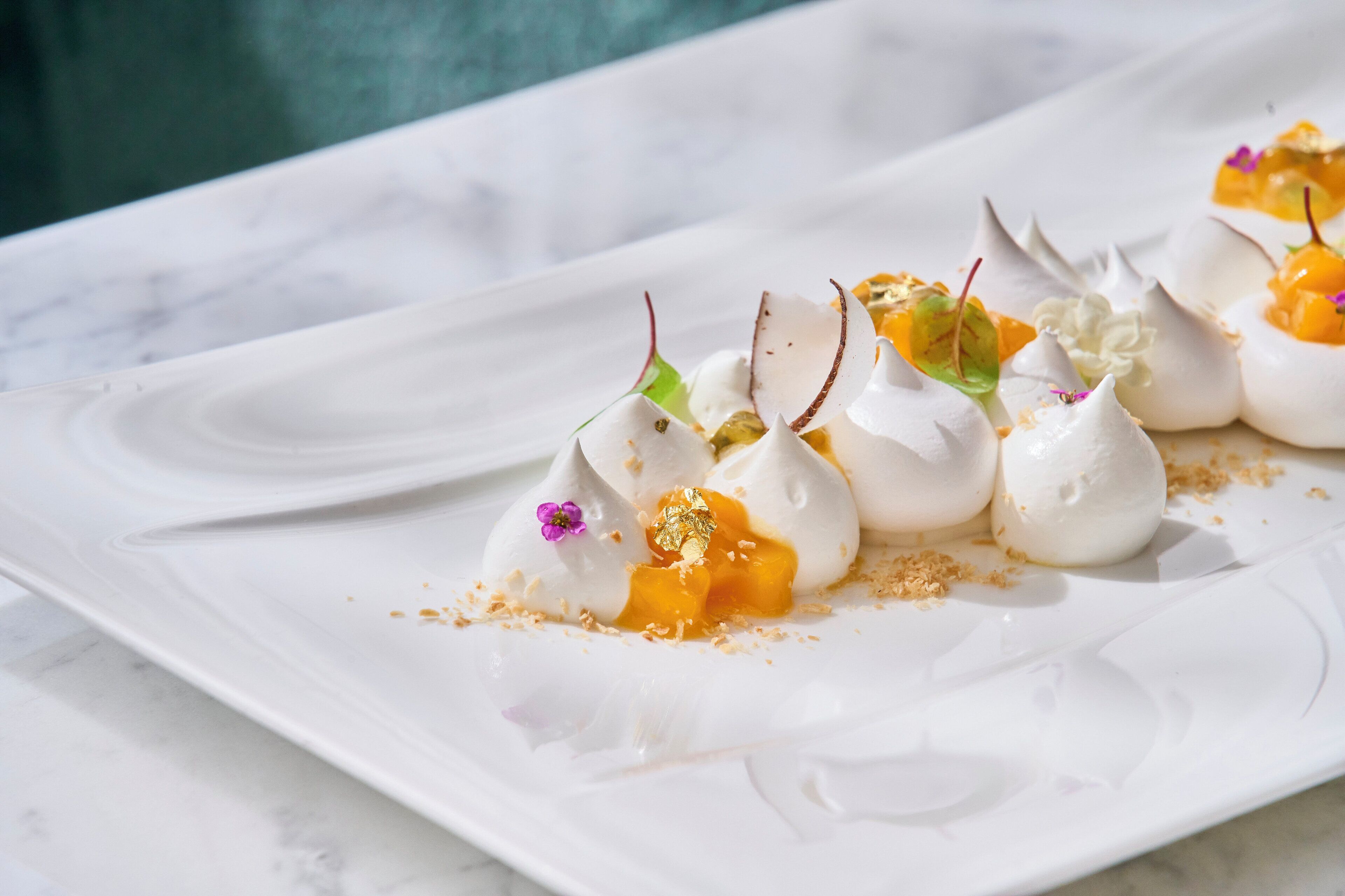 Un plato blanco con un postre de picos parecidos al merengue, cubierto de mango y flores comestibles.