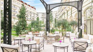 A covered outdoor seating area with tables and chairs, surrounded by greenery and overlooking a residential building.