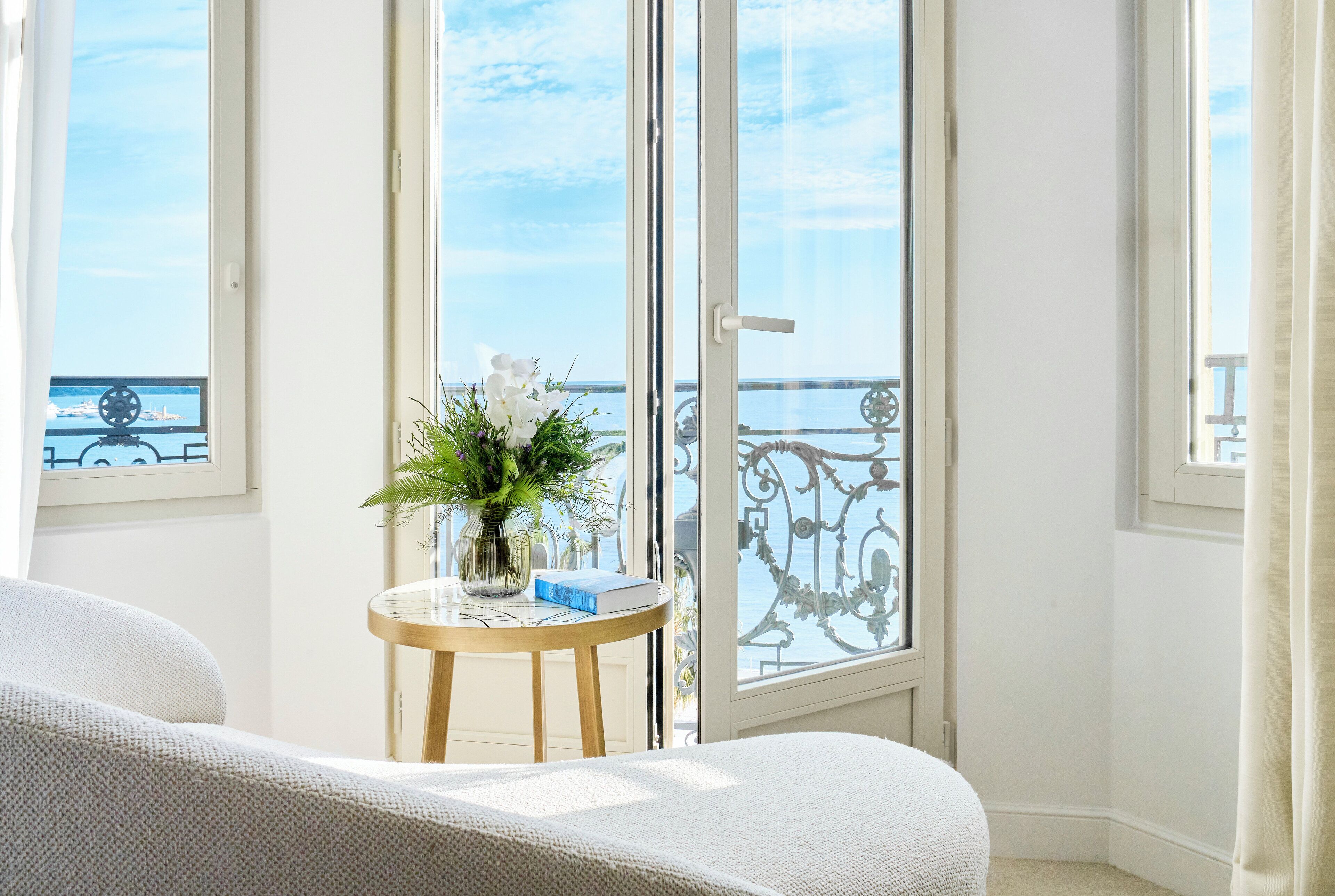 A room with a large window overlooking a balcony with a view of the sea. There is a small round table with a vase of flowers and a book.