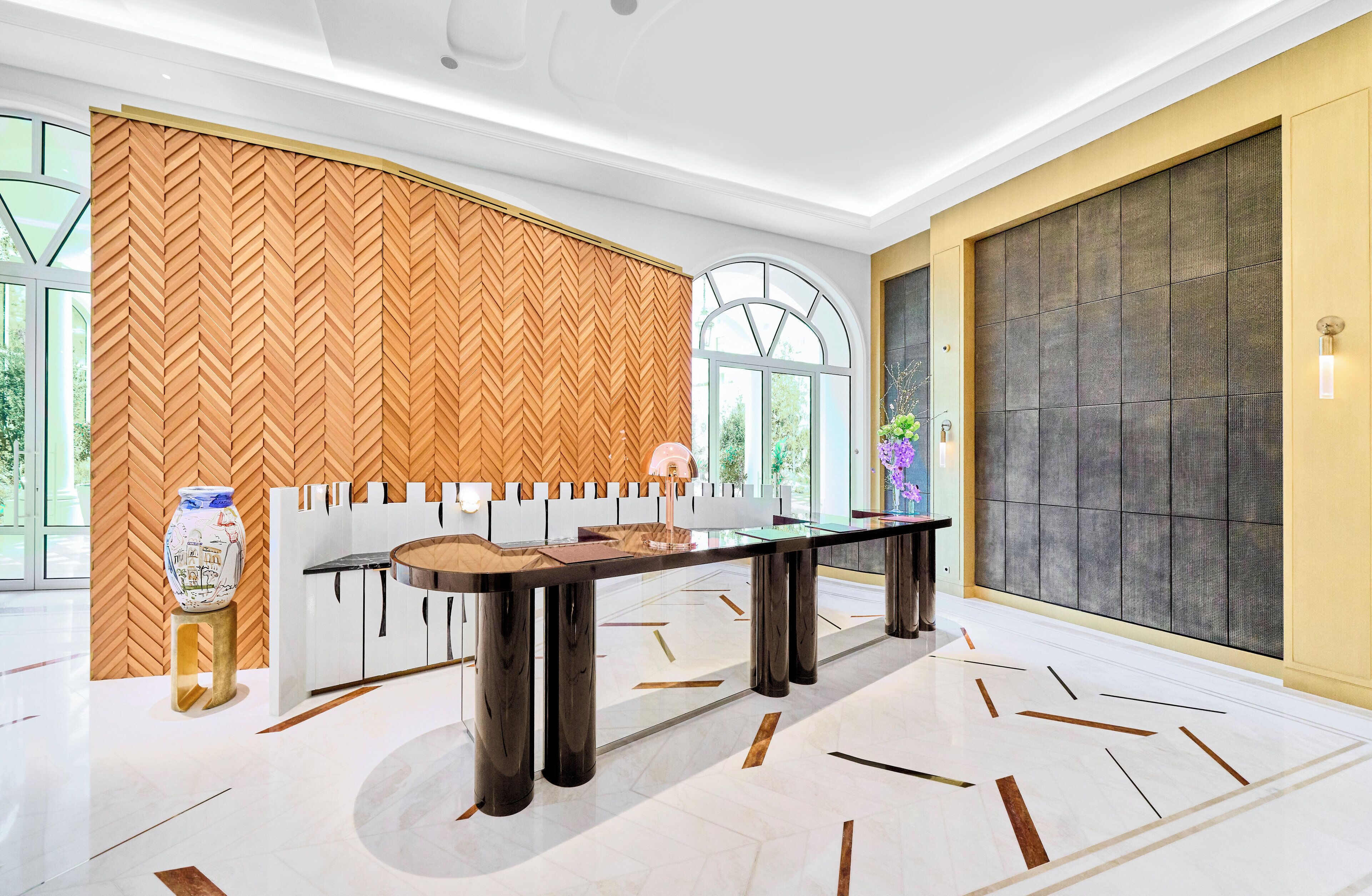 A modern hotel lobby with a large wooden herringbone wall, a glass-top table, and a large window with a view of greenery.
