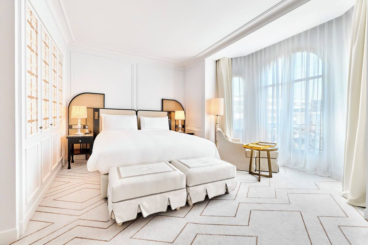 A spacious bedroom with a large bed, two bedside tables with lamps, a small round table, and a large window with sheer curtains.