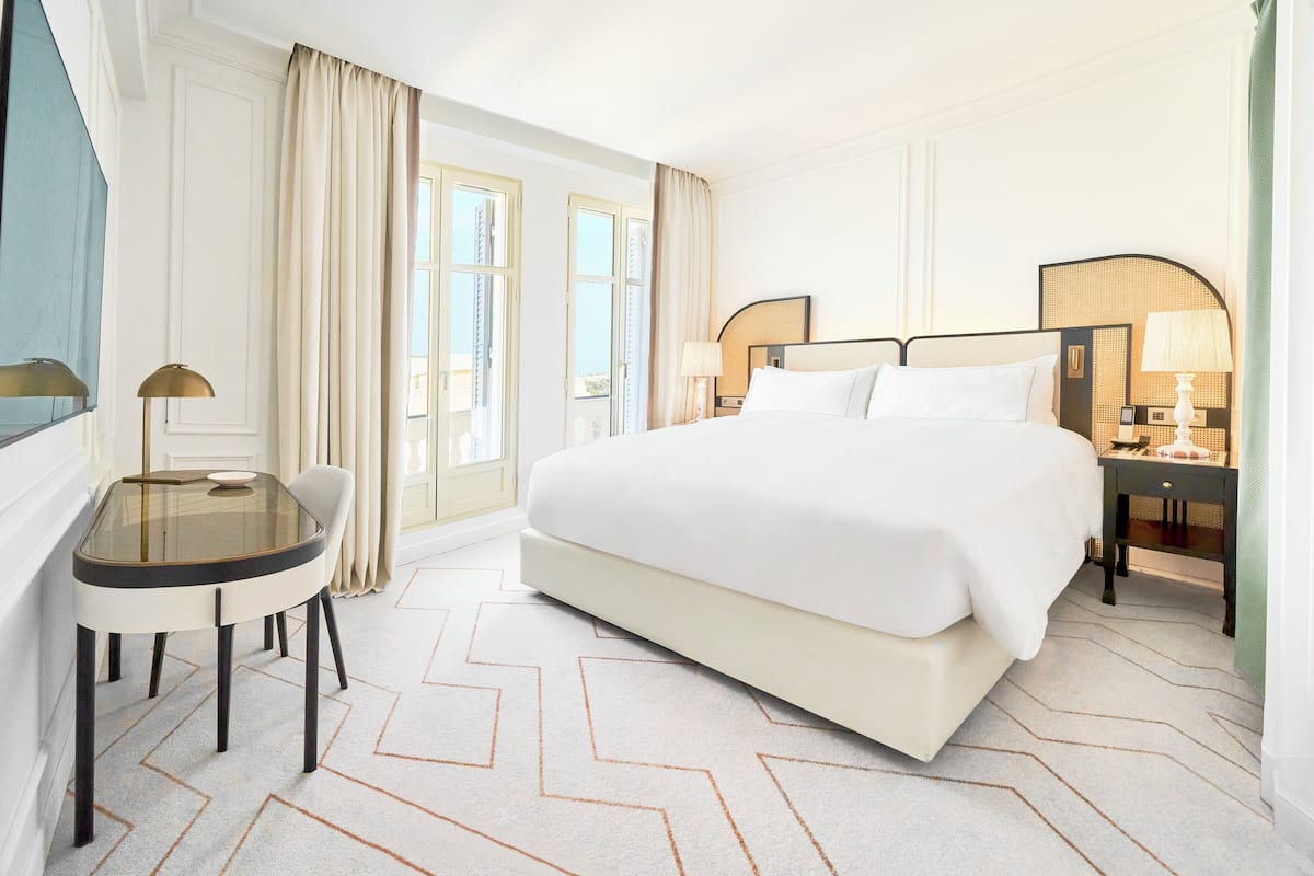 A hotel room with a large bed, a desk with a lamp, a bedside table with a lamp, and a large window with curtains.