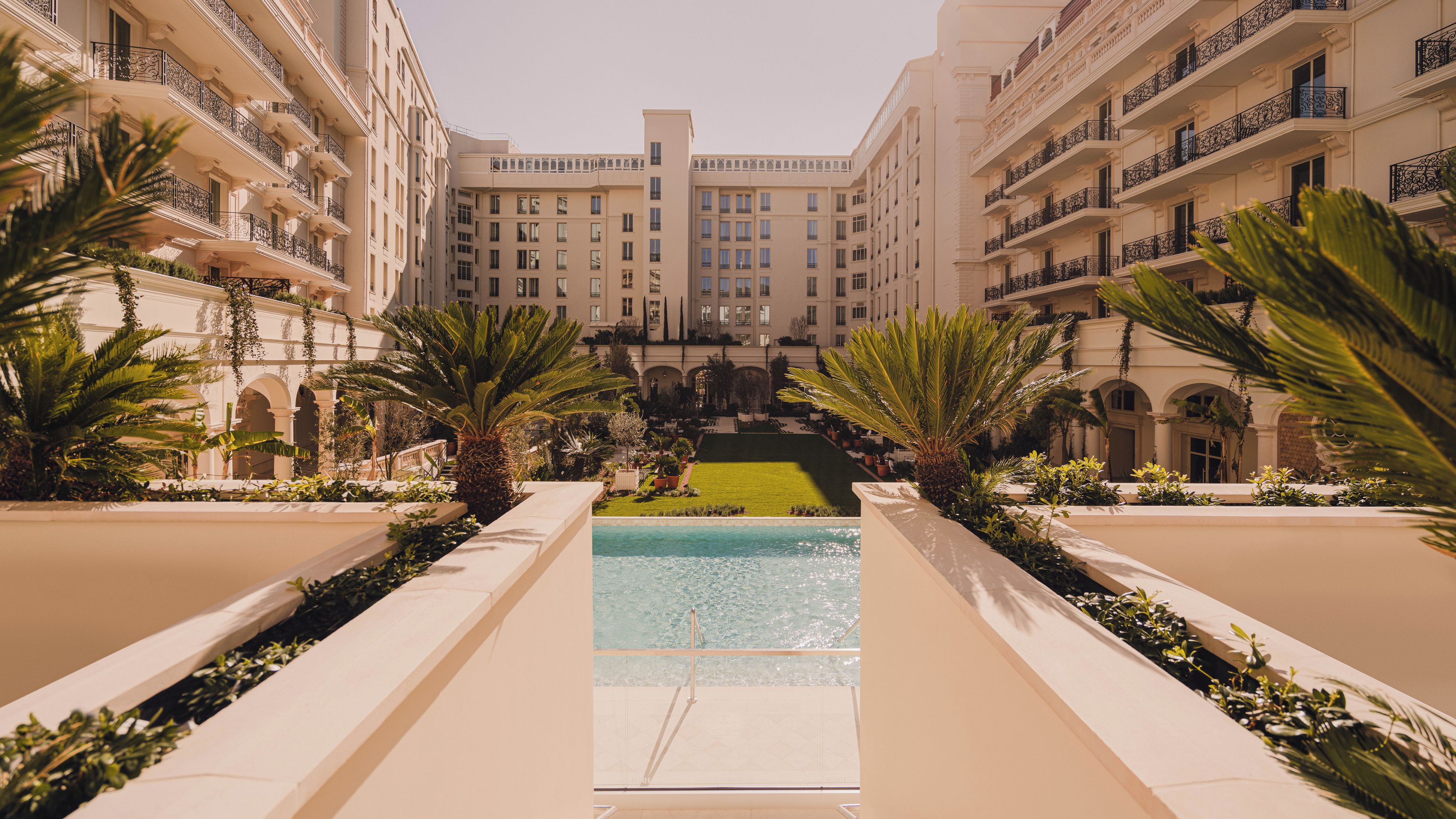 Pátio de hotel com piscina, palmeiras e um edifício central com vários andares e varandas.