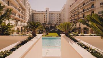 Pátio de hotel com piscina, palmeiras e um edifício central com vários andares e varandas.