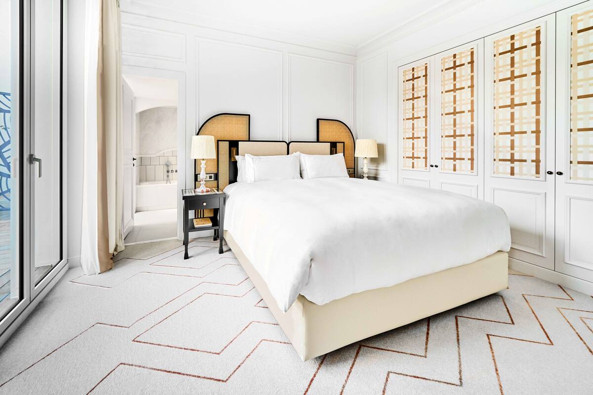 A spacious bedroom with a large bed, a nightstand, a lamp, and a wardrobe with a geometric pattern.