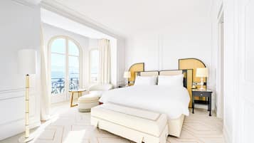 A spacious bedroom with a large bed, two bedside tables, a chair, and a view of the sea through a large window.