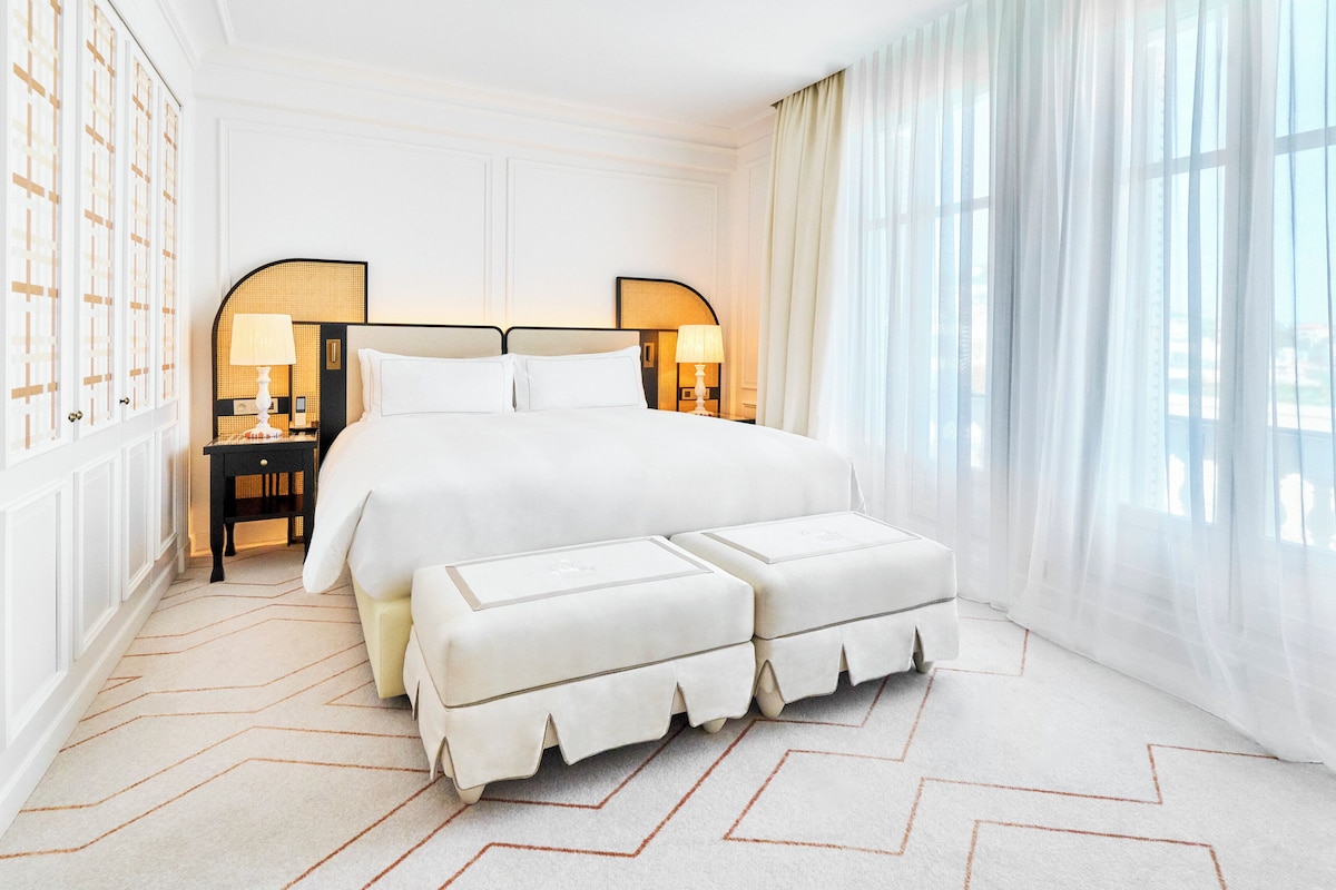 A spacious bedroom with a large bed, two bedside tables with lamps, a white upholstered bench, and a view of the city through sheer curtains.