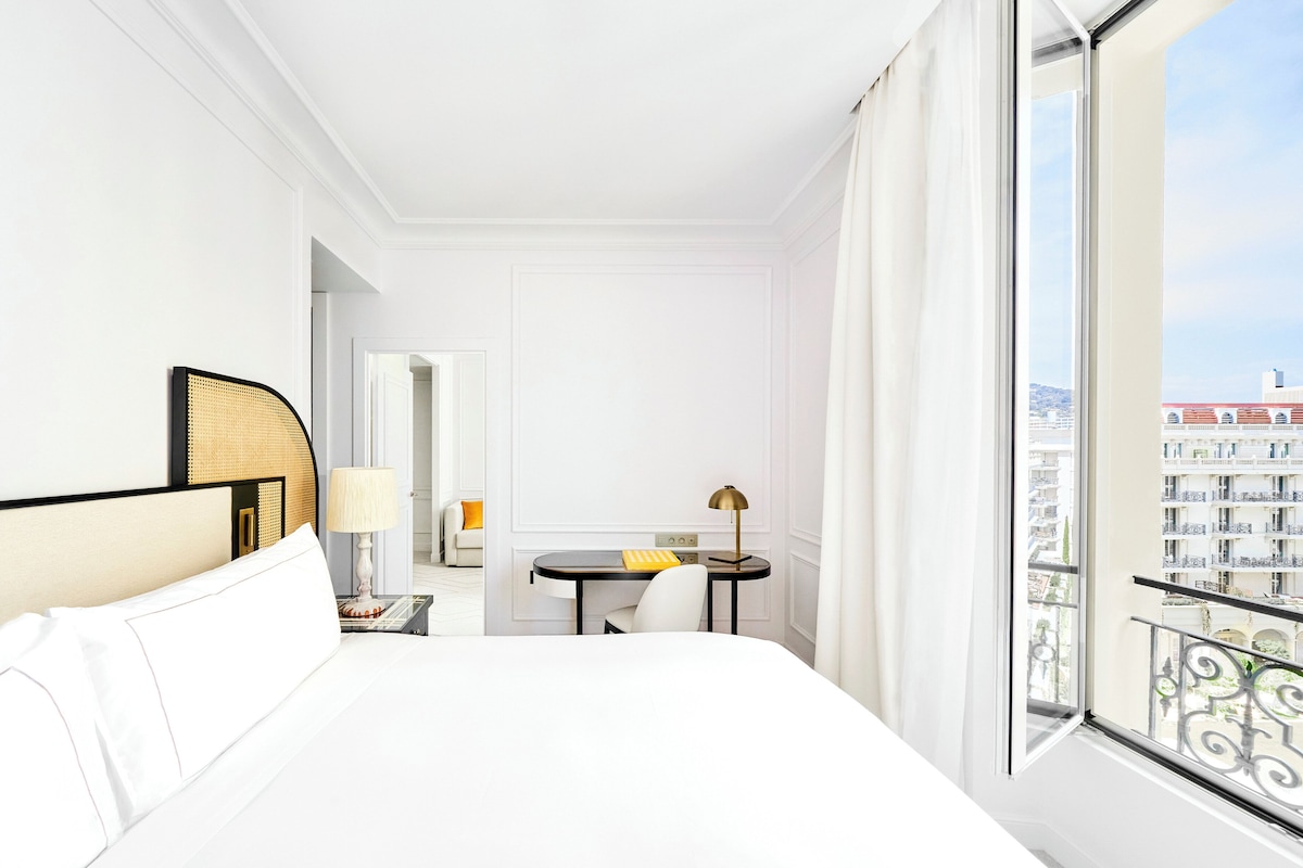 A hotel room with a bed, a desk, a chair, and a view of the city through large windows.