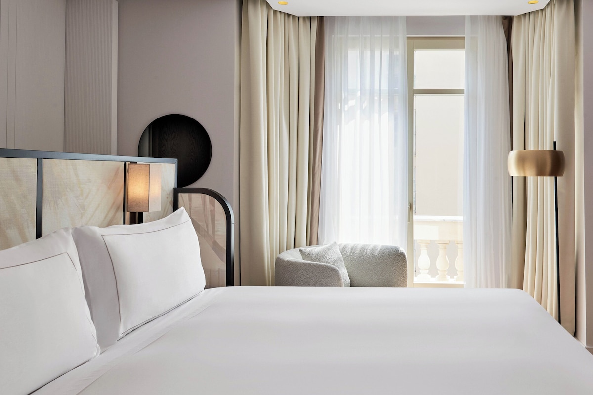 A hotel room with a large bed, a bedside table, a chair, and a window with curtains.