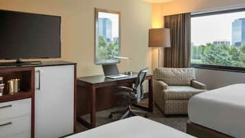 A hotel room with a TV, a desk with a laptop, a chair, a bed, and a window with a city view.