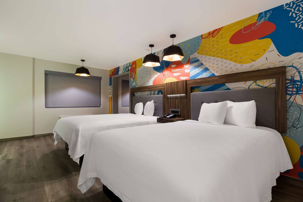 A hotel room with two beds, a large abstract mural on the wall, and pendant lights.