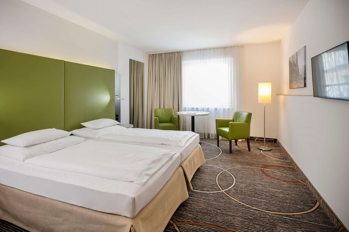 A hotel room with two beds, a green headboard, a small table, a green chair, a lamp, and a painting on the wall.
