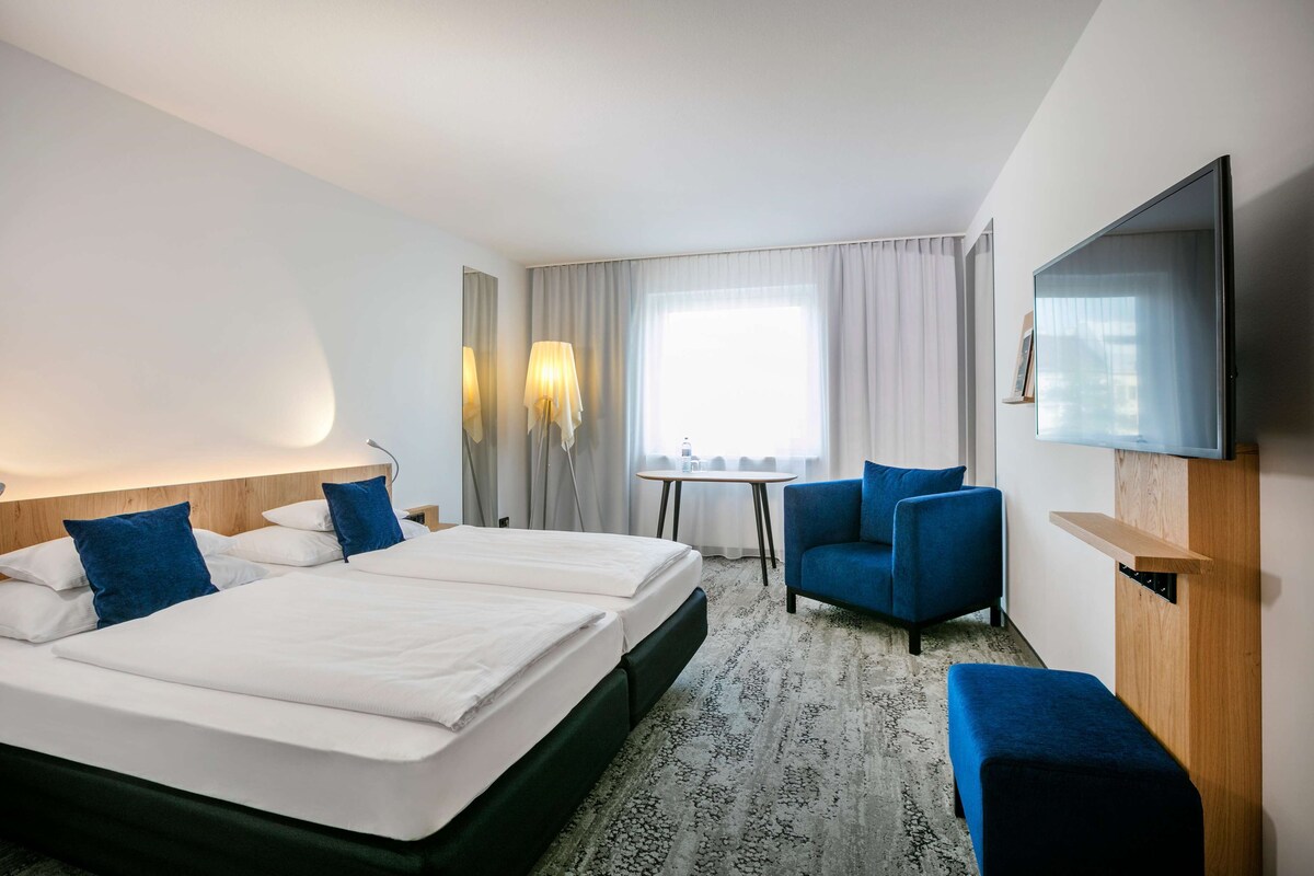 A hotel room with a large bed, a desk, a chair, a TV, and a window with curtains.