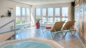 Un área de piscina interior moderna con dos sillones de mimbre, una planta en maceta roja y grandes ventanales con vistas a la ciudad.