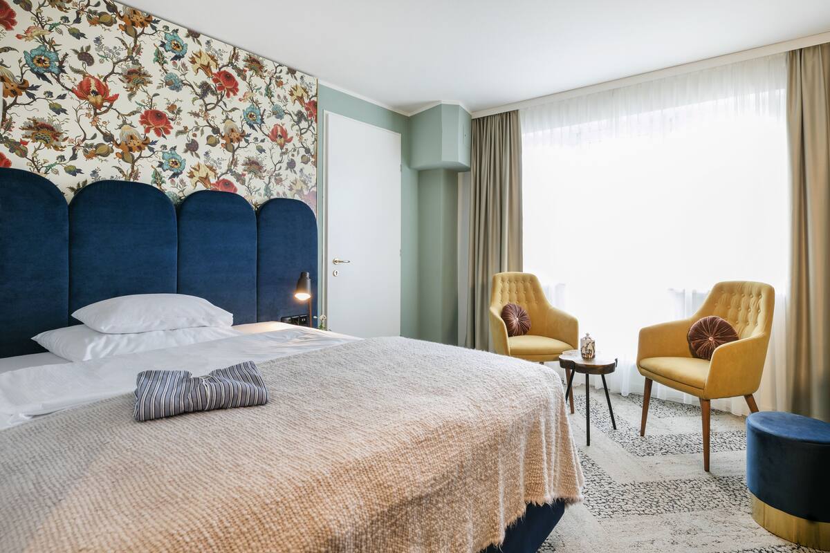 Une chambre d’hôtel avec un grand lit, deux fauteuils, une petite table et une fenêtre avec des rideaux.