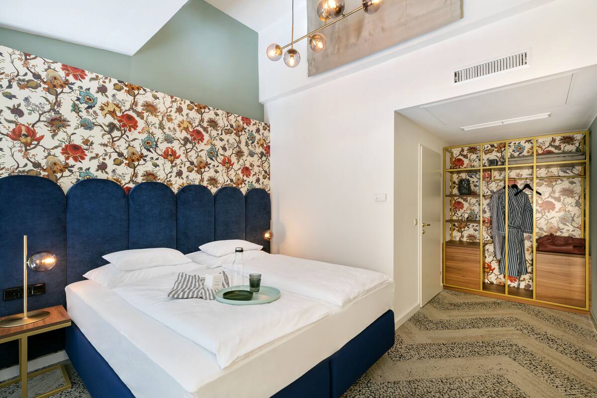 Une chambre moderne avec un grand lit, une tête de lit bleue, un mur orné d’un motif floral et une armoire au décor floral.