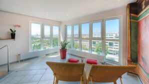 Un balcón espacioso con ventanas amplias, sillones de mimbre, una planta en maceta roja y vistas a la ciudad.