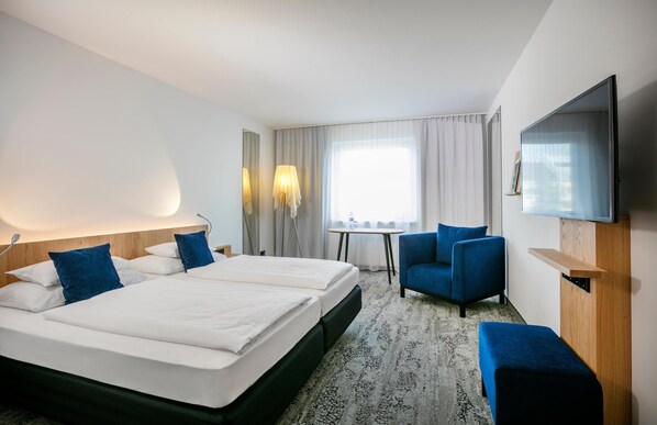 A hotel room with a large bed, a desk, a chair, a TV, and a window with curtains.