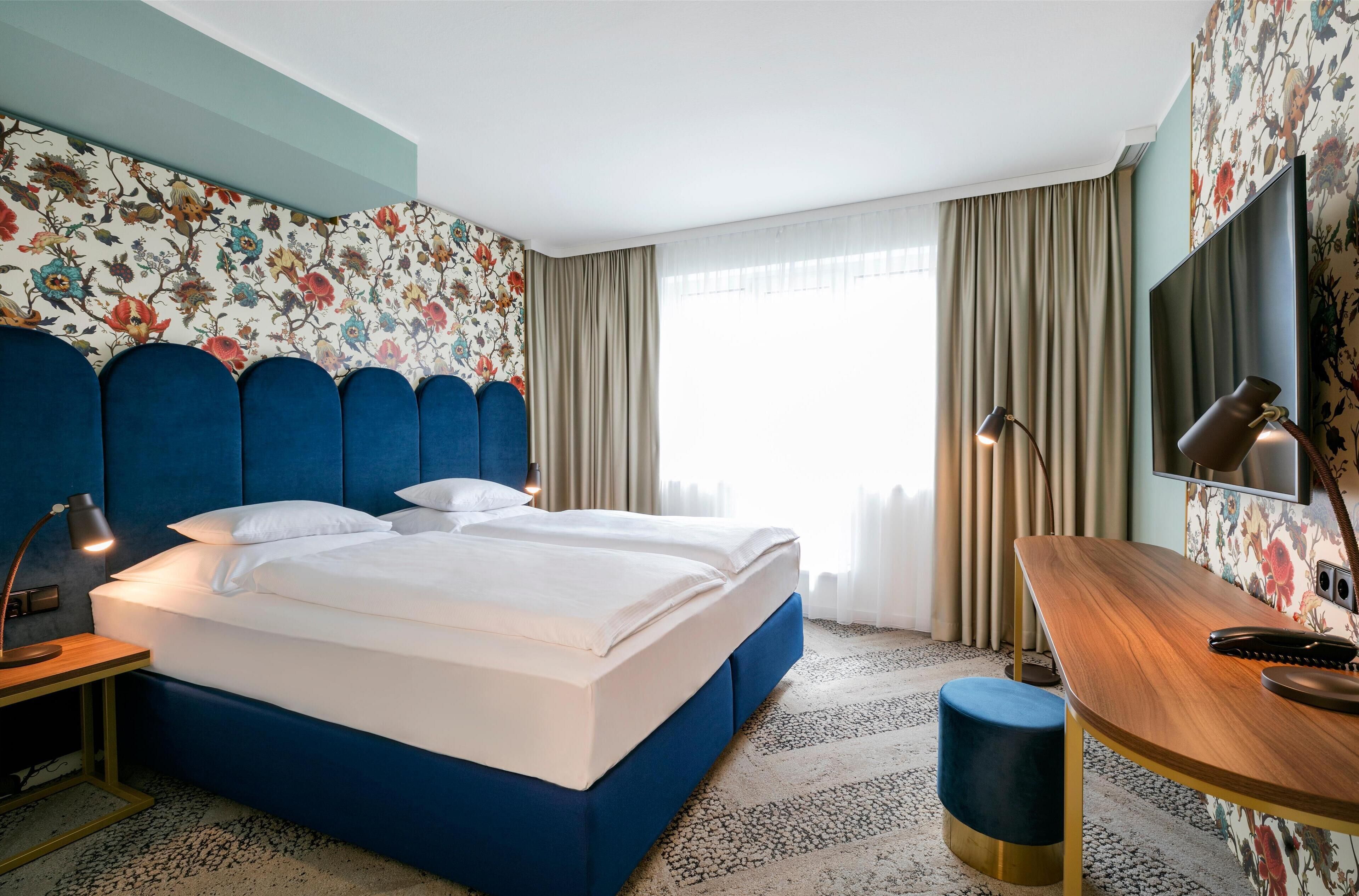 Ein Hotelzimmer mit einem großen Bett, einem Schreibtisch, einem Fernseher und floralem Wandpapier.