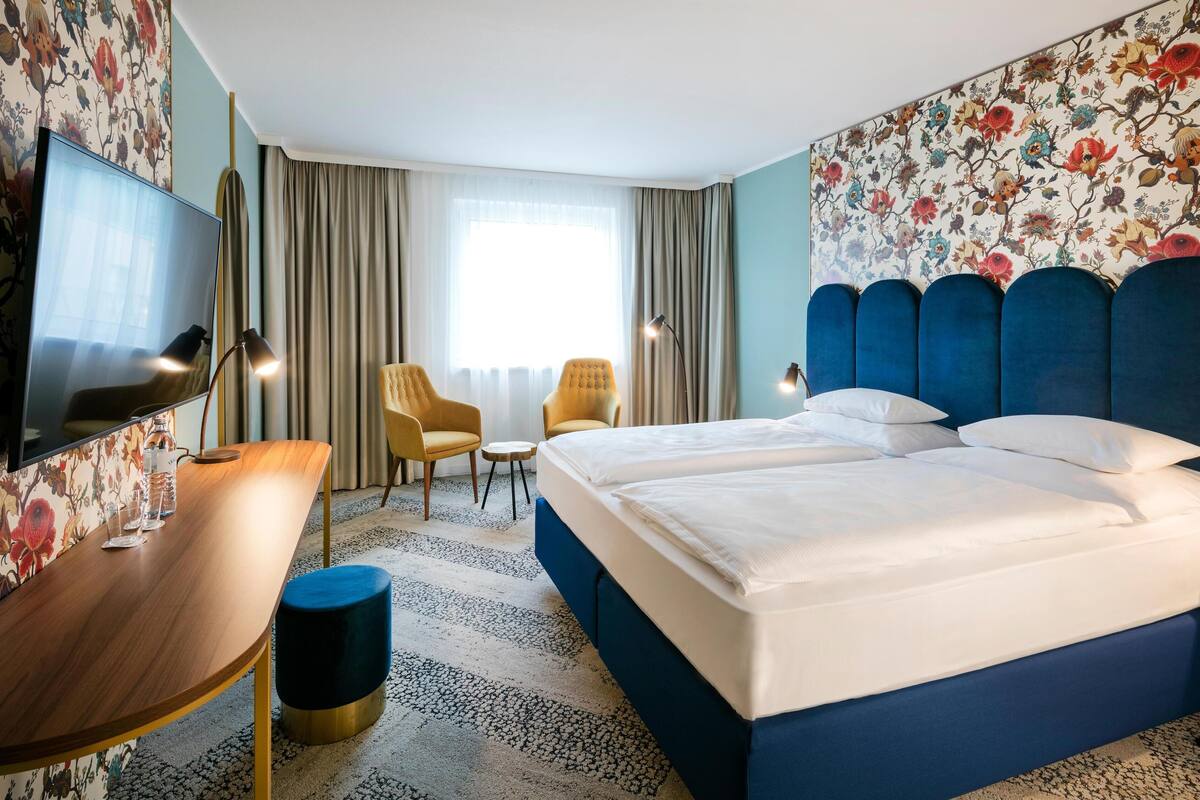Une chambre d’hôtel avec un grand lit, un bureau, deux chaises, une télévision et un papier peint à motifs floraux.