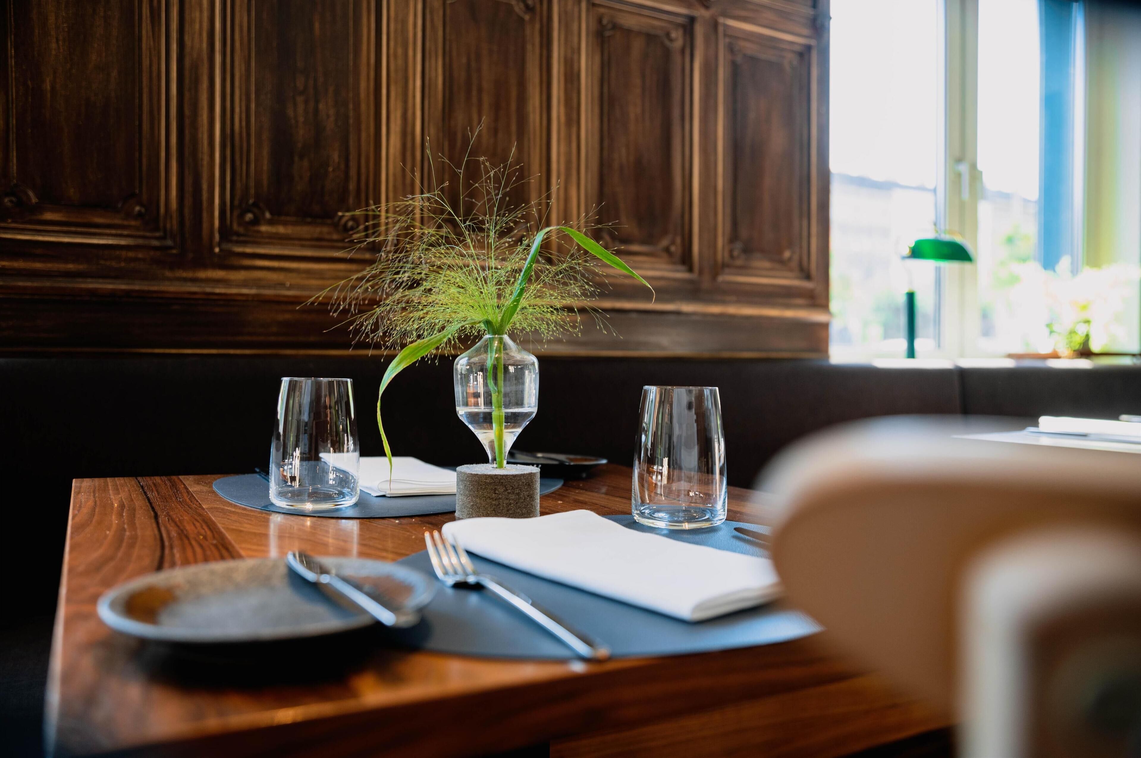 Una mesa de comedor con un jarrón de plantas verdes, dos vasos vacíos, un tenedor y un cuchillo.