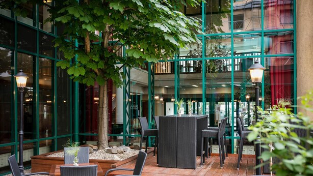 Un patio al aire libre con mobiliario de mimbre, un árbol y un edificio de vidrio al fondo.