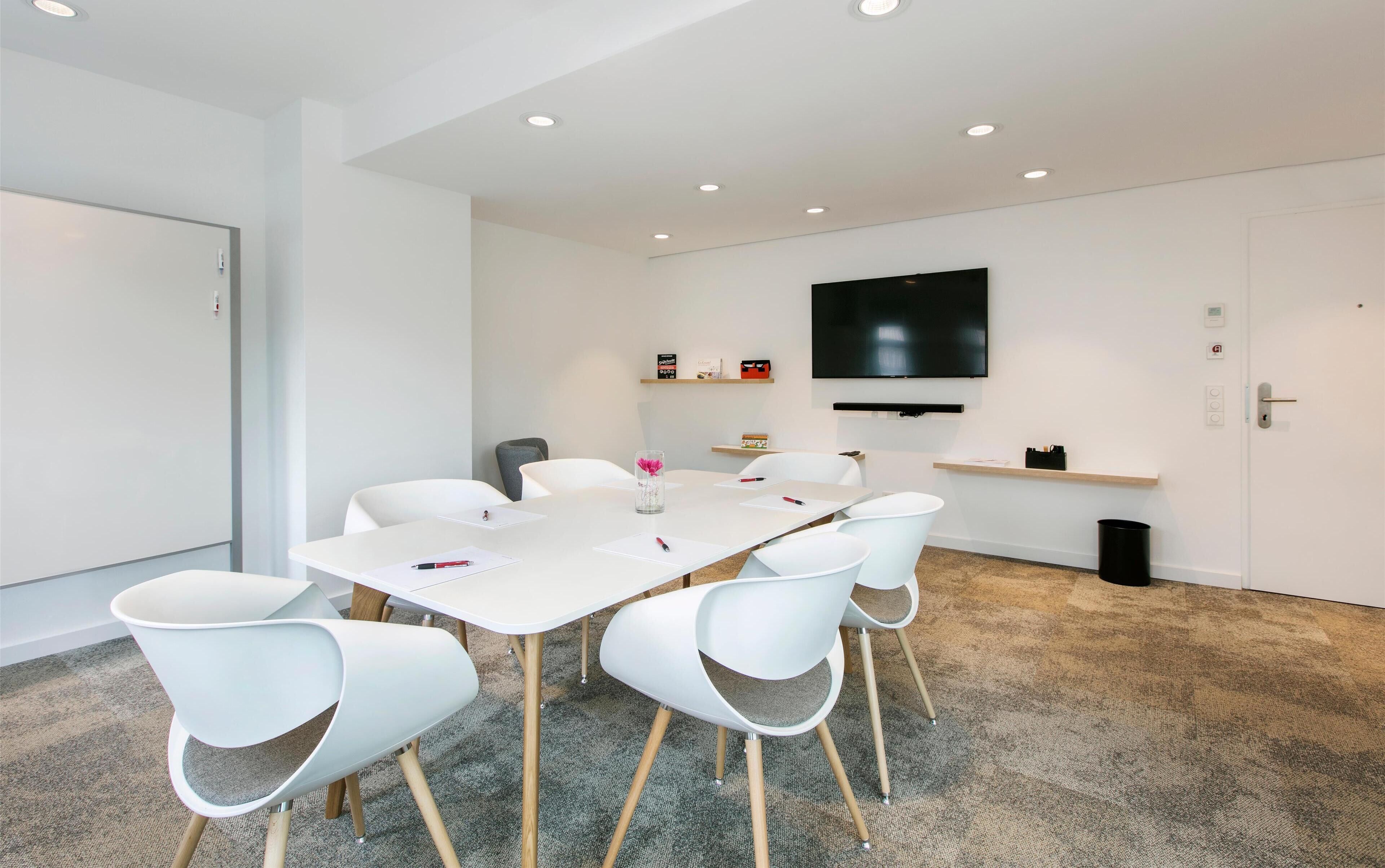 A conference room with a white table, white chairs, a mounted TV, and a carpeted floor.