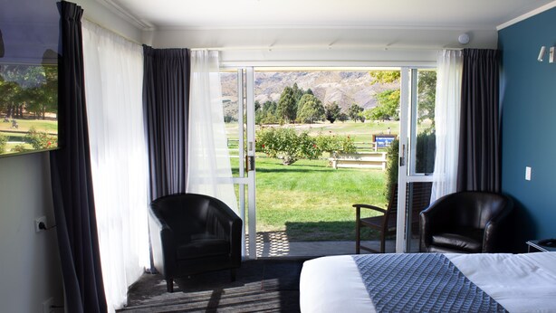 A hotel room with a large bed, two chairs, a view of a green landscape through a sliding door, and a carpeted floor.