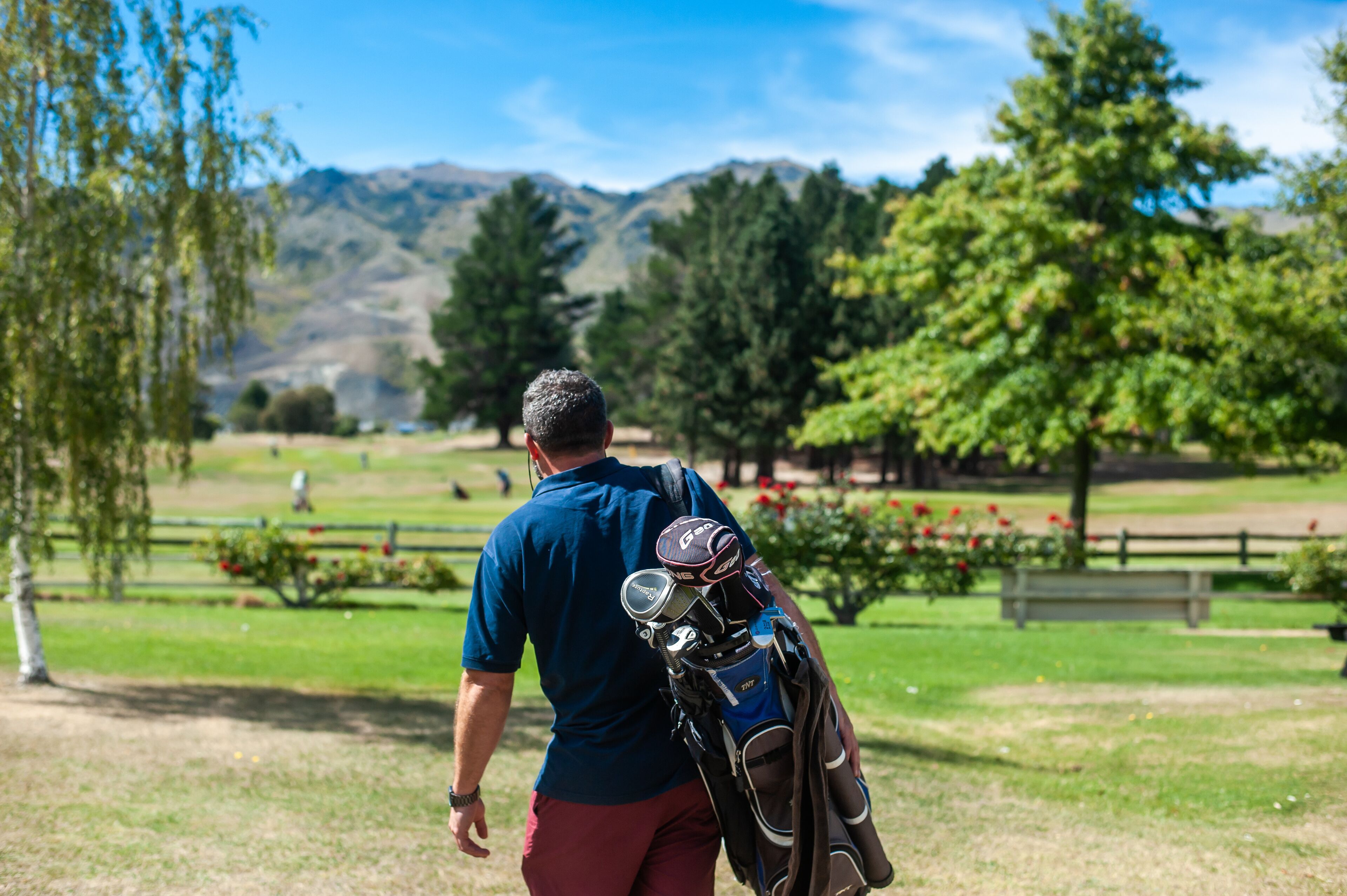 A man carrying golf bags on a golf course with trees and mountains in the background.