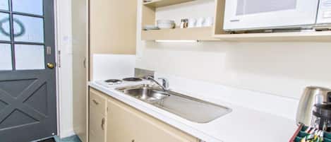 A compact kitchen with a white countertop, a stainless steel sink, and a microwave above a shelf with dishes.
