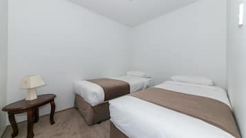 Una habitación con dos camas individuales, una mesita de madera con una lámpara y una pared blanca lisa.