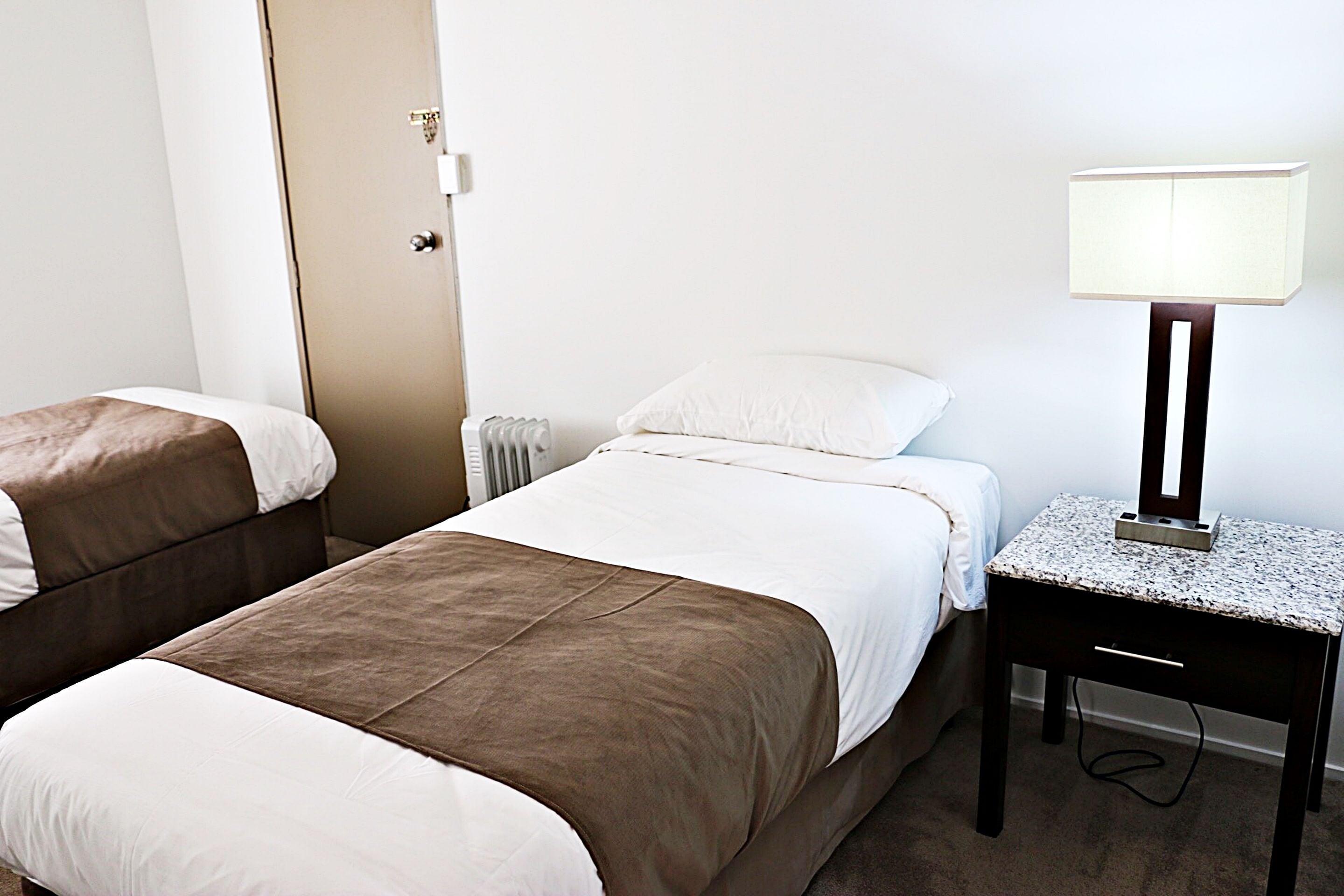 A hotel room with two beds, a nightstand with a lamp, and a radiator.
