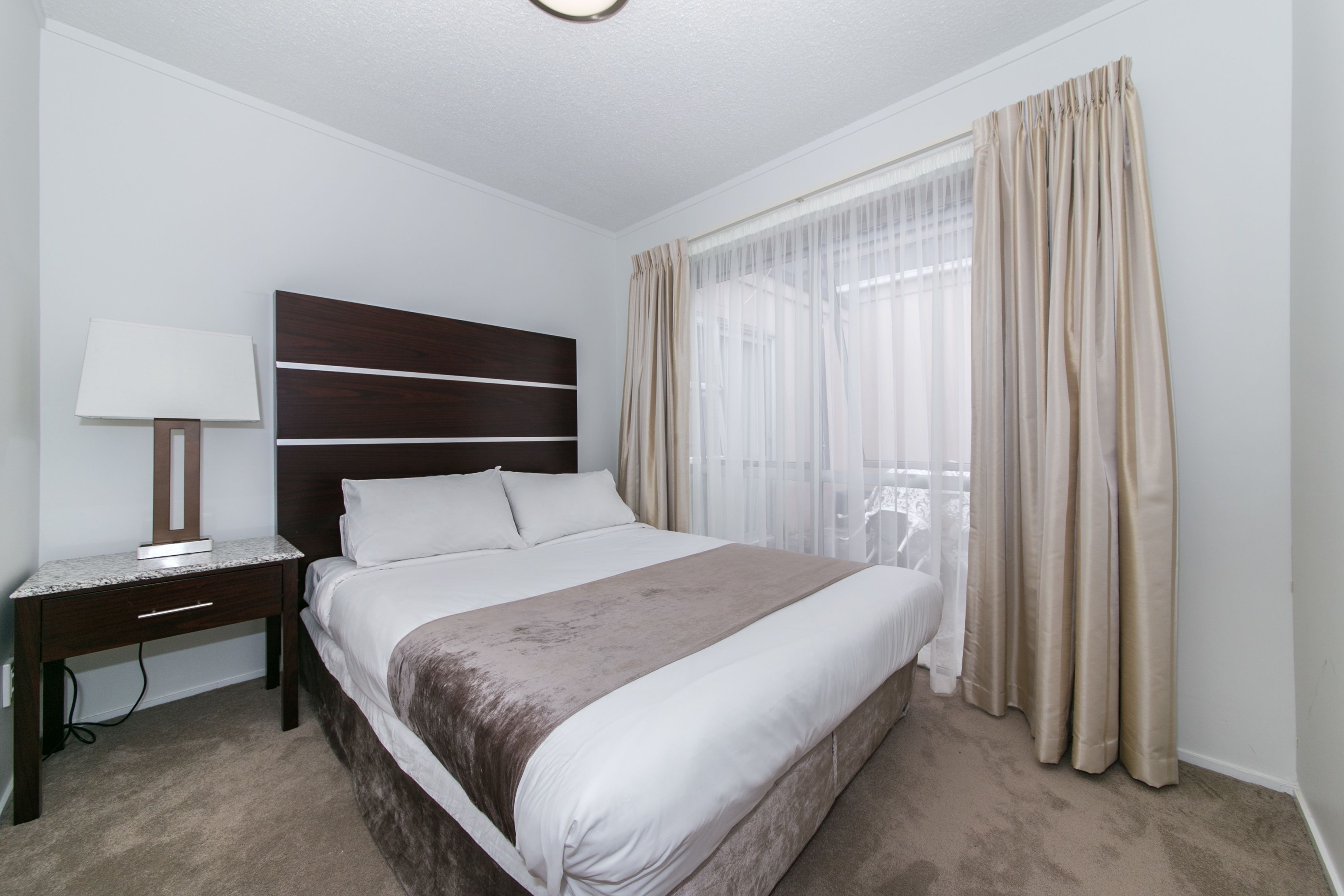 Une chambre d’hôtel avec un grand lit, une table de chevet avec une lampe et des rideaux légers à la fenêtre.