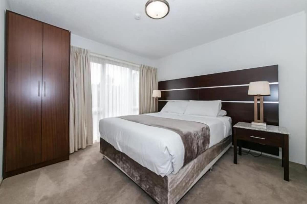 A hotel room with a large bed, a wardrobe, a bedside table with a lamp, and a window with curtains.