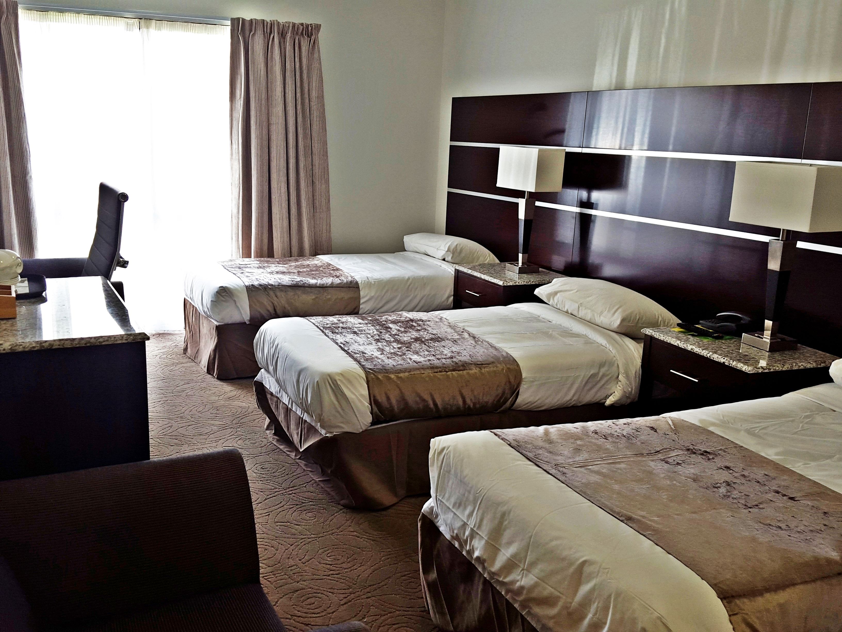 A hotel room with two beds, a desk, a chair, a nightstand with a lamp, and a window with curtains.