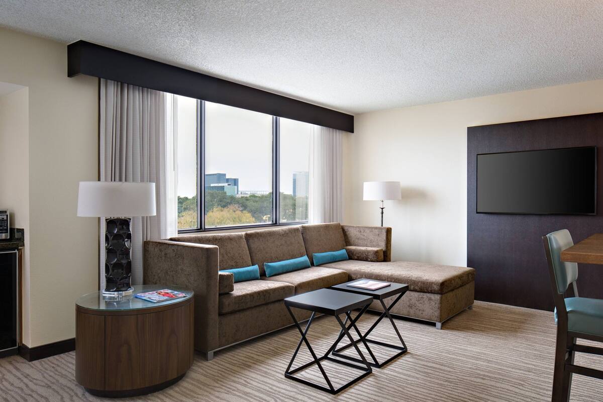 A modern hotel room with a sectional sofa, a small table, a TV mounted on the wall, and a window with a city view.