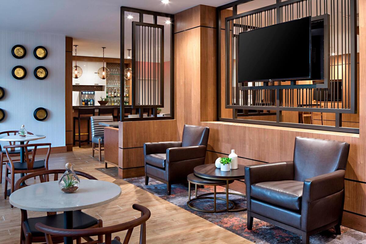 A modern hotel lobby with leather armchairs, a round table, a bar area, and a television mounted on the wall.