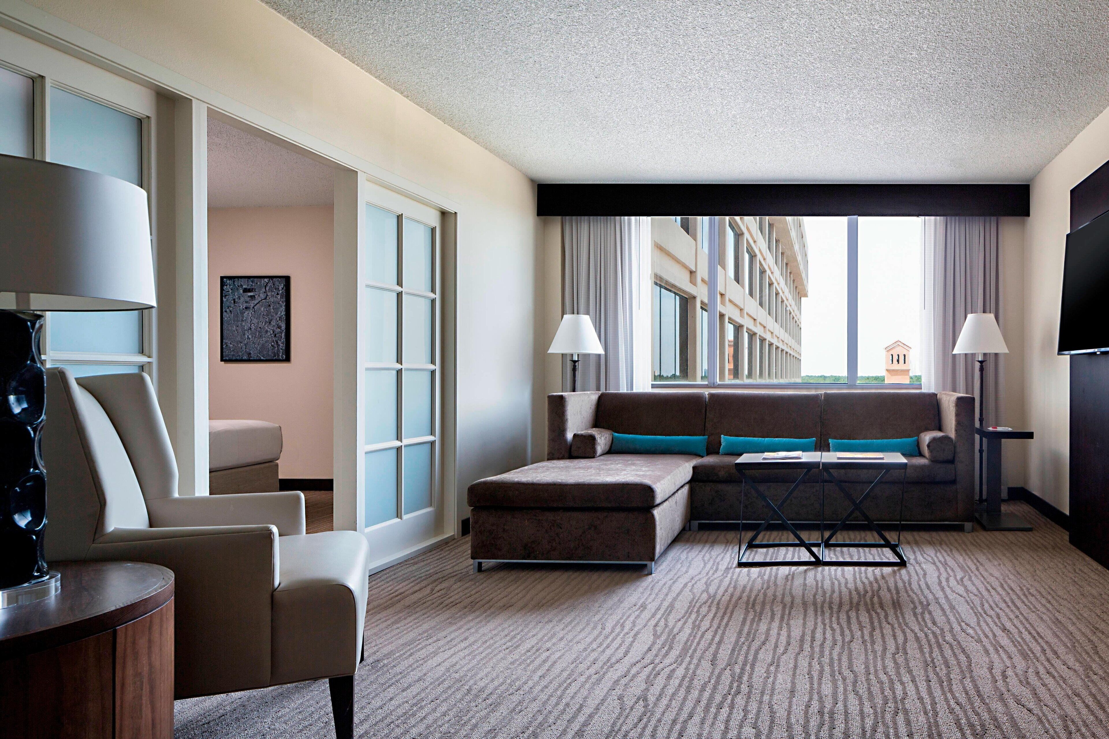A hotel room with a sofa, armchair, and a small table. There is a window with curtains, a flat-screen TV, and a lamp.