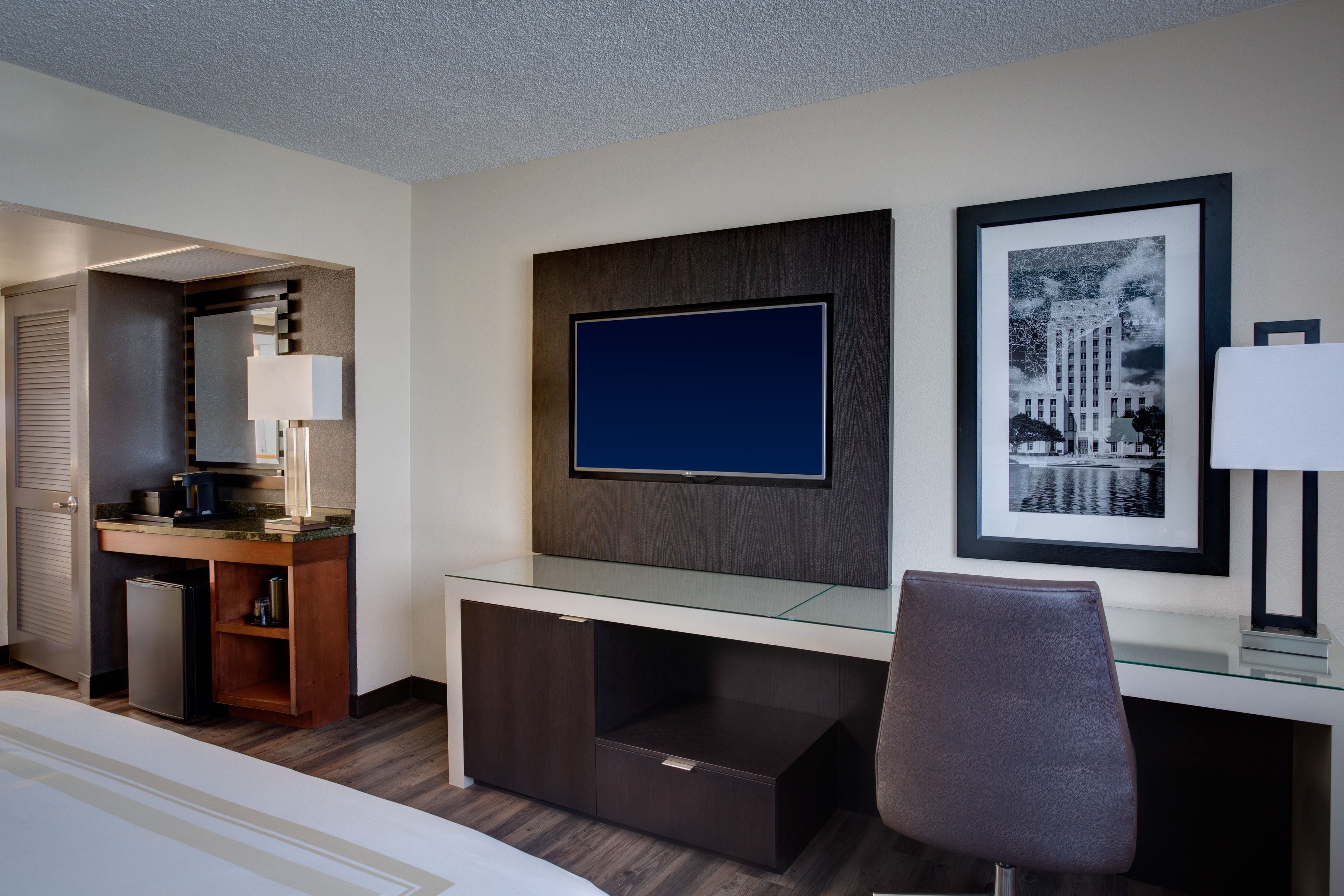 A modern hotel room with a glass desk, a flat-screen TV, a framed cityscape picture, and a compact kitchenette.