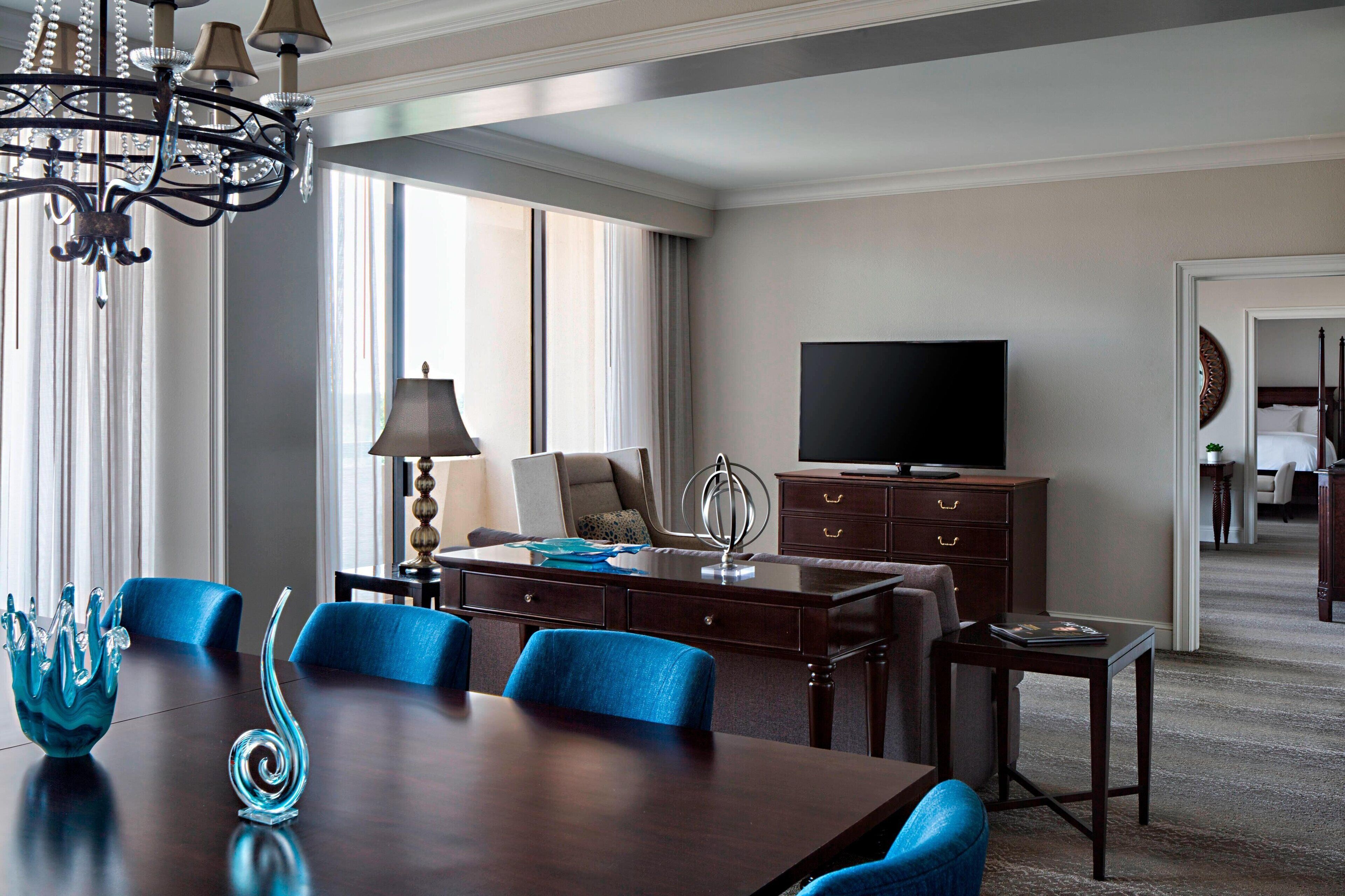 Een hotelkamer met een zithoek, bestaande uit een donkerhouten tafel, blauwe stoelen met bekleding en decoratieve glazen vazen. Een televisie is aan de muur bevestigd, en er is een bureau met lades te zien.