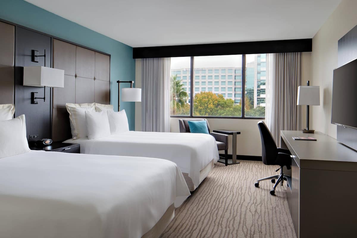 A hotel room with two beds, a desk, a chair, and a TV.