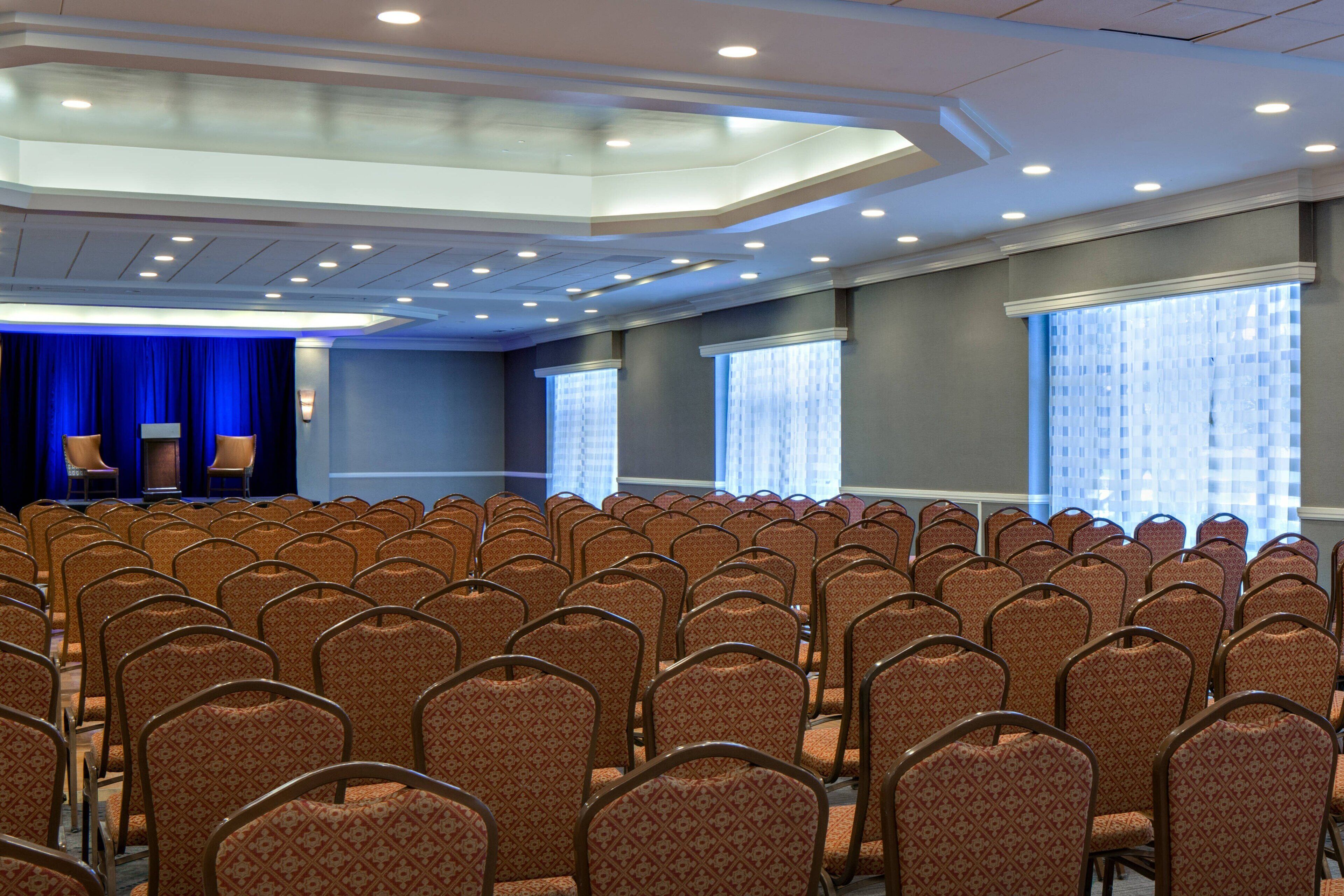 Un salón de conferencias con filas de sillas, un escenario con dos sillas y ventanas amplias con persianas.